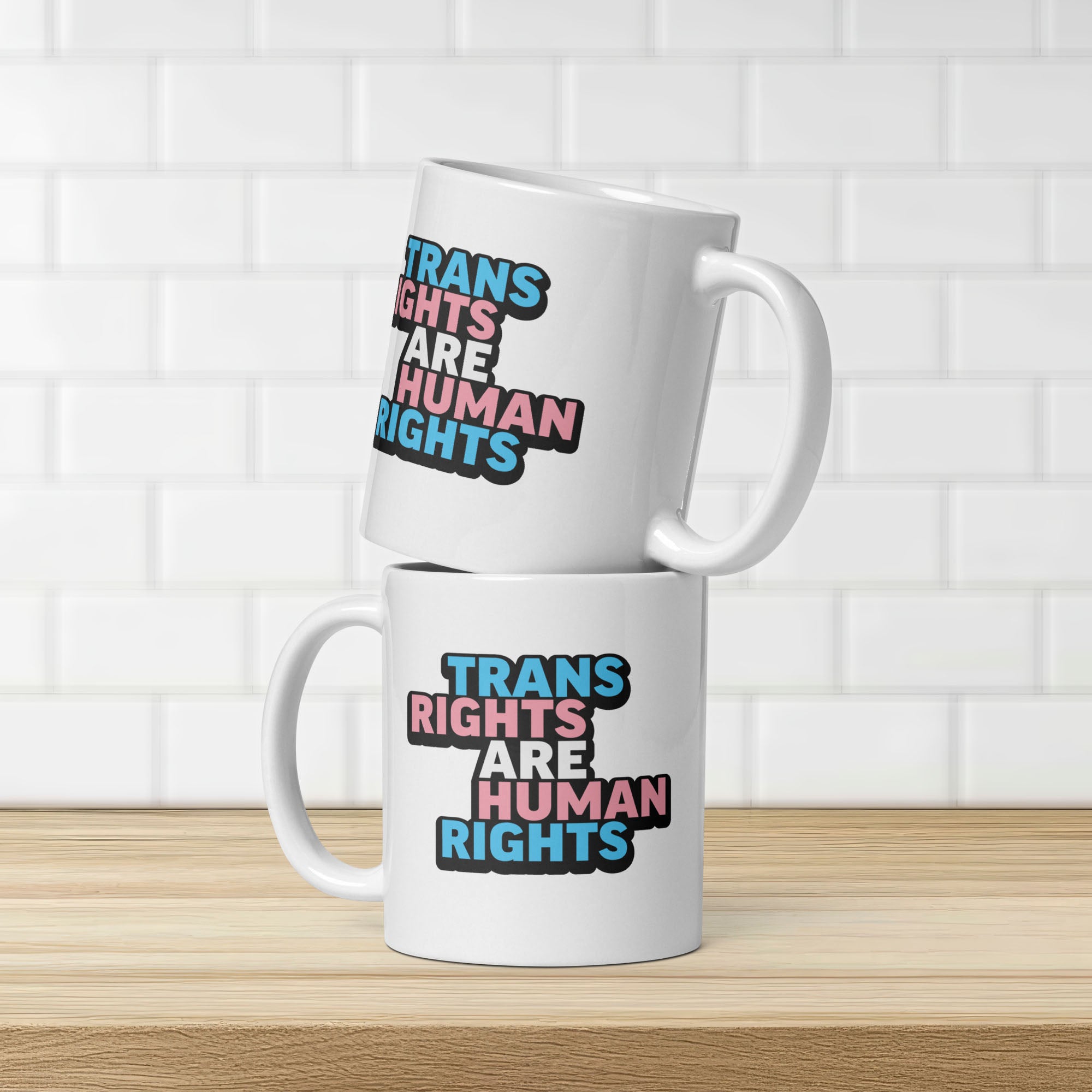 Two stacked mugs. Each mug has bold text “Trans Rights Are Human Rights” in blue, pink, and black colors representing the transgender pride flag. Shows that same design is printed on both sides of the mug.