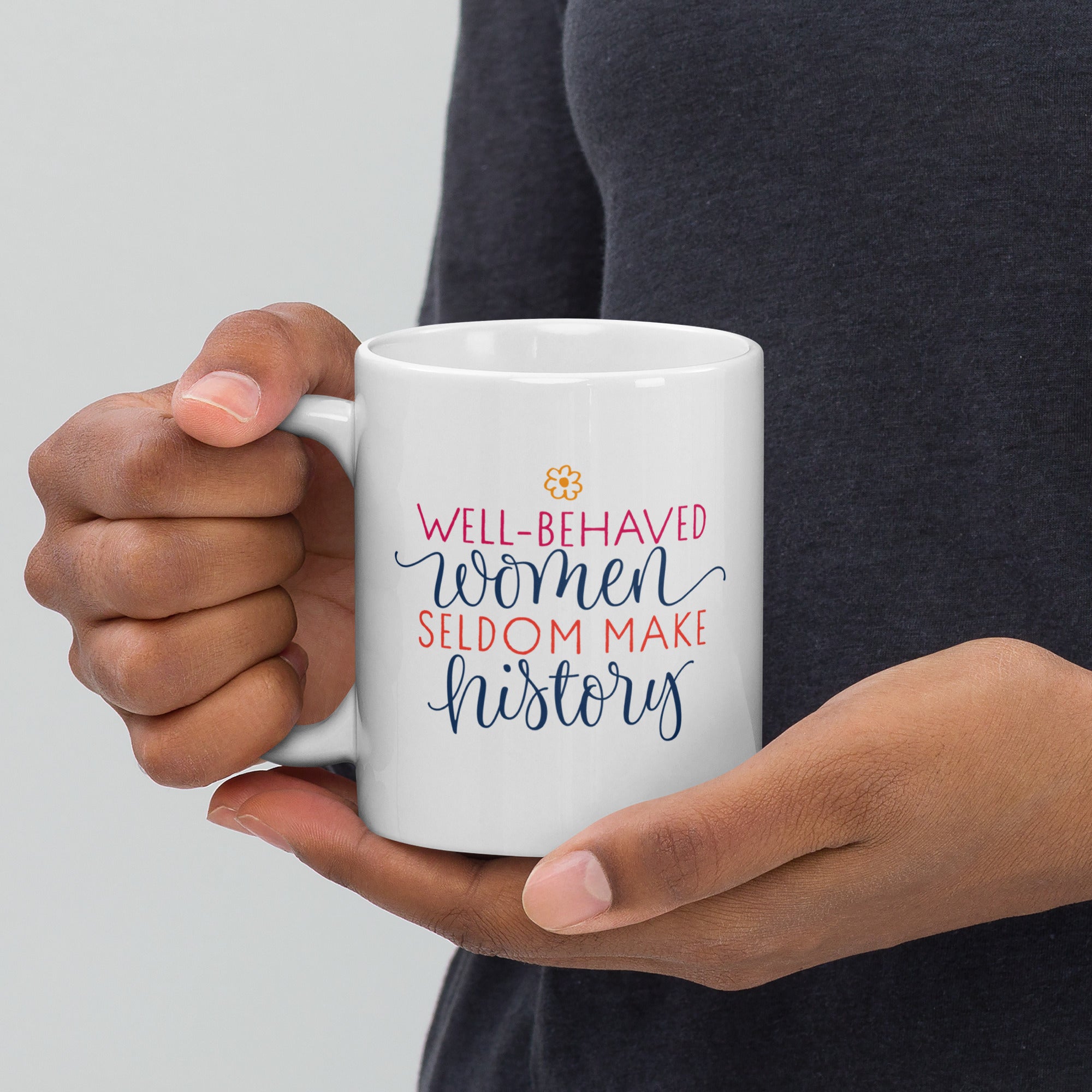 Person holding a white mug with colorful text that reads "well-behaved women seldom make history" against a neutral background