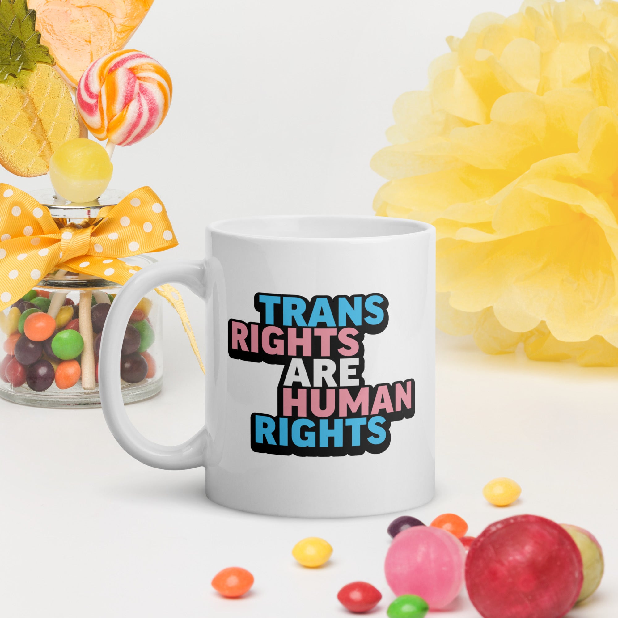 White ceramic mug with bold text “Trans Rights Are Human Rights” in blue, pink, and black colors representing the transgender pride flag, on a table surrounded by brightly colored candy.