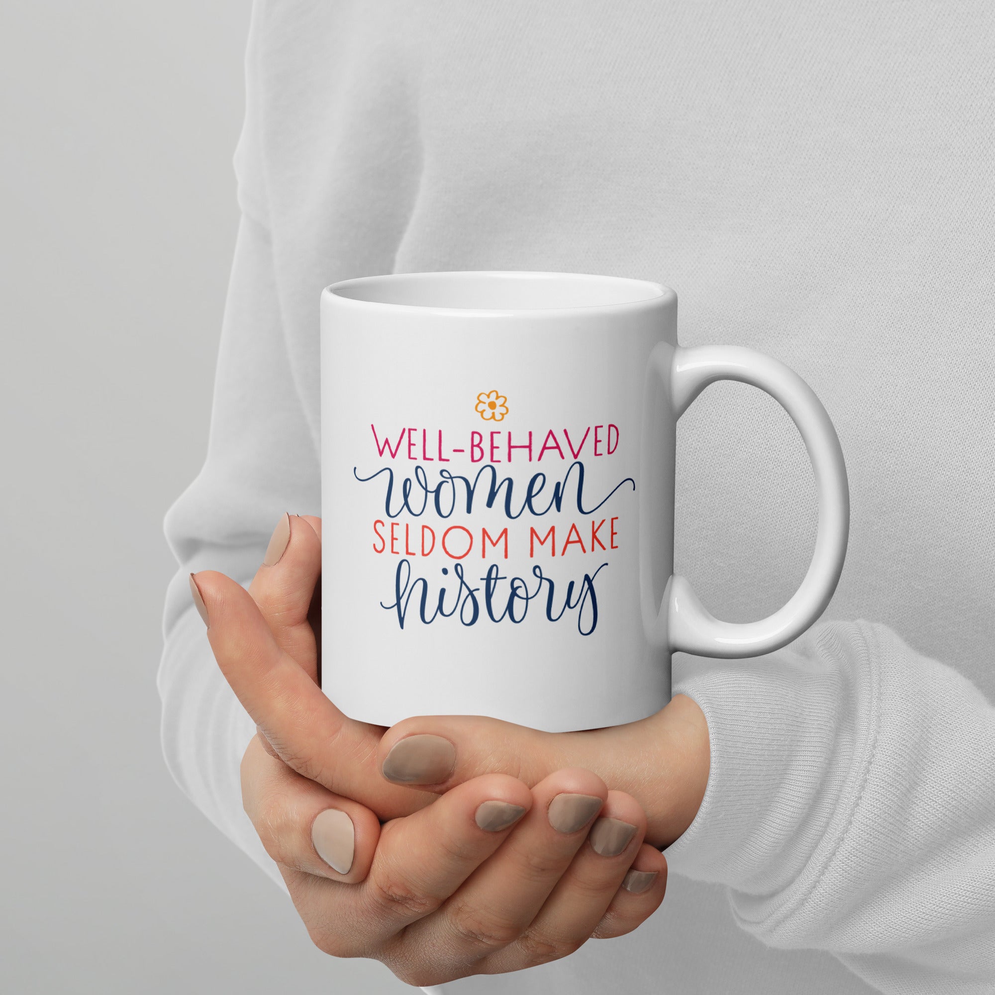 White mug with 'Well-behaved women seldom make history' text held by a person against a gray background.