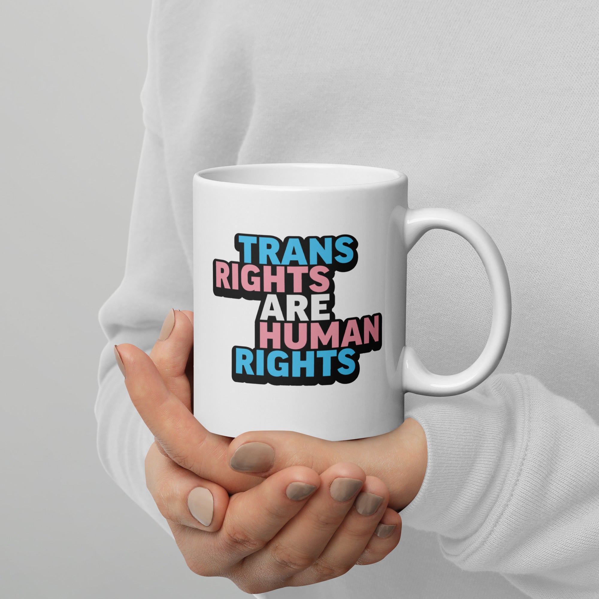 Person holding white ceramic mug with bold text “Trans Rights Are Human Rights” in blue, pink, and black colors representing the transgender pride flag, with handle on the right.