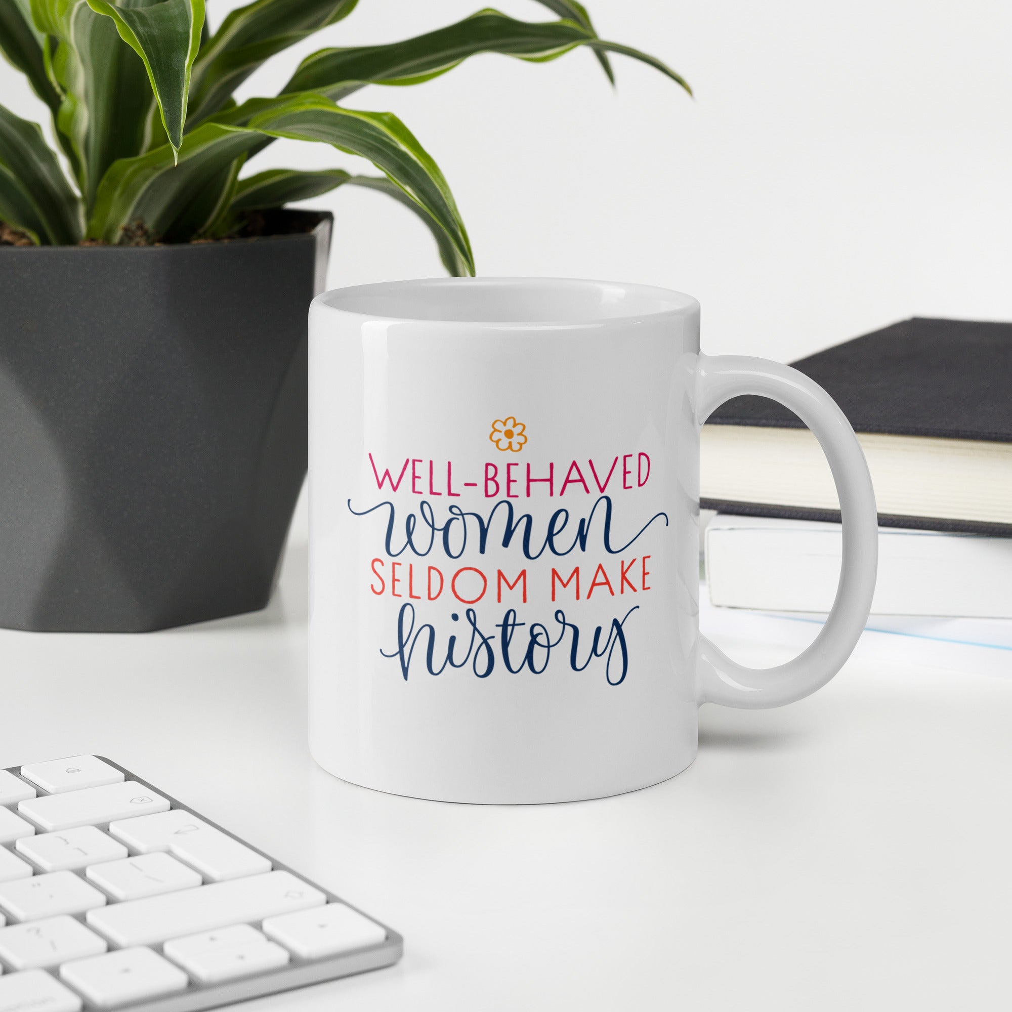 White mug with motivational quote that reads "well-behaved women seldom make history" on a desk with keyboard, plant, and notebook.