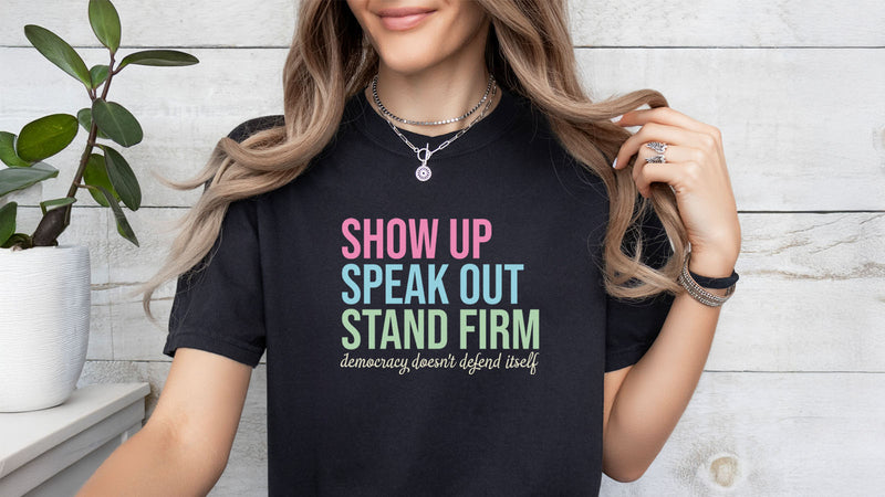 Female model wearing black t-shirt that reads "show up, speak out, stand firm, democracy doesn't defend itself."