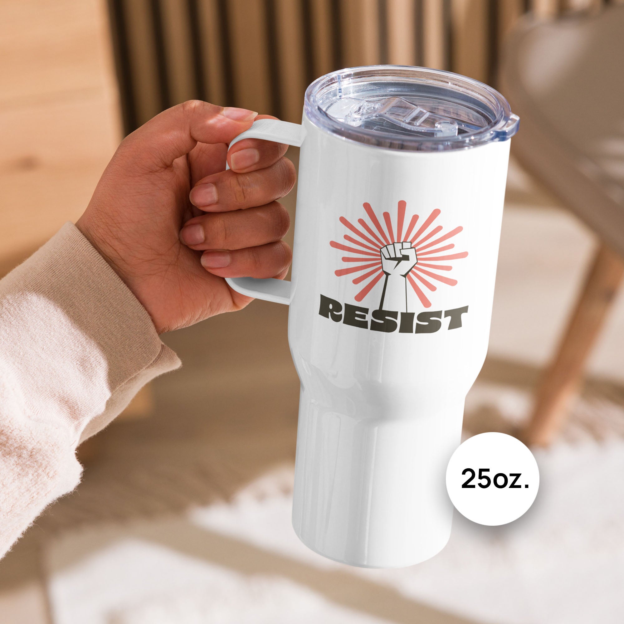 25 ounce white stainless steel tumbler with handle, with graphic text "resist" below a raised fist and coral color burst, held by a person on an outdoor deck.