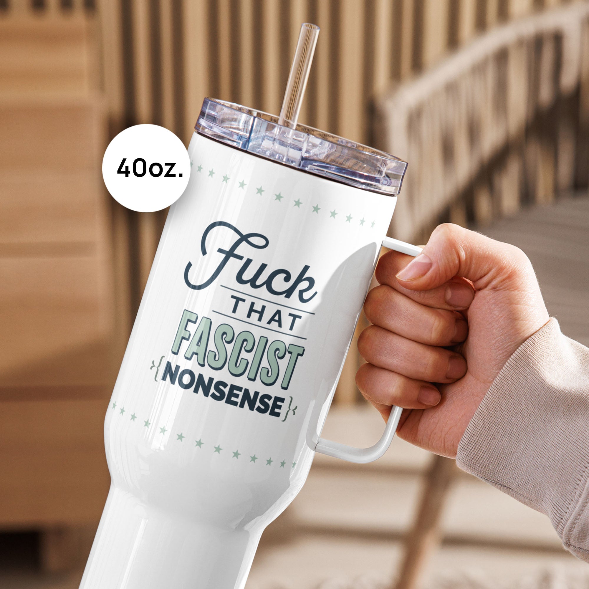 Person holding 40 ounce white insulated travel tumblers with handle featuring the bold quote “Fuck That Fascist Nonsense” in blue and mint green text, on a counter in a kitchen, on an outdoor deck.