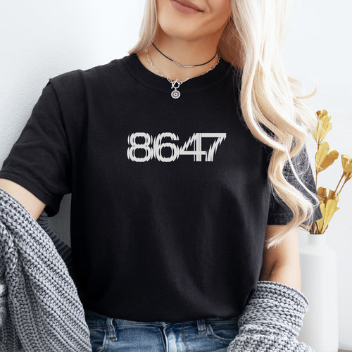 Female model wearing black unisex t-shirt featuring the number “8647” in bold, clean typography with glass block effect, referencing Trump impeachment.