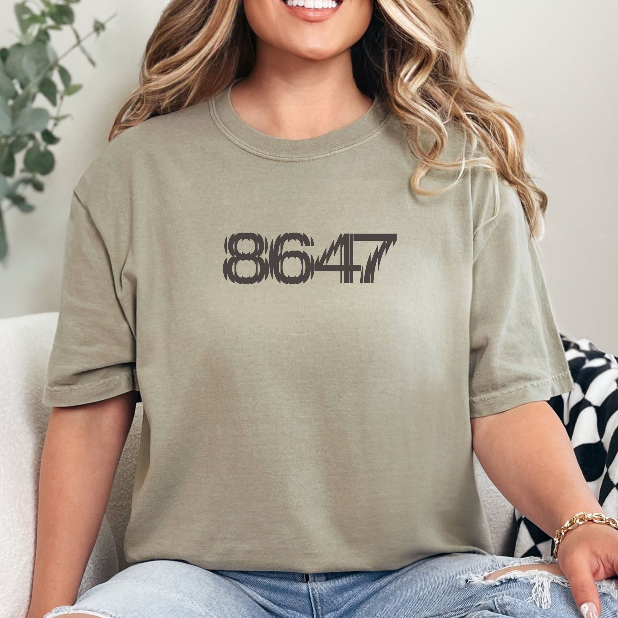 Female model wearing khaki unisex t-shirt featuring the number “8647” in bold, clean typography with glass block effect, referencing Trump impeachment.