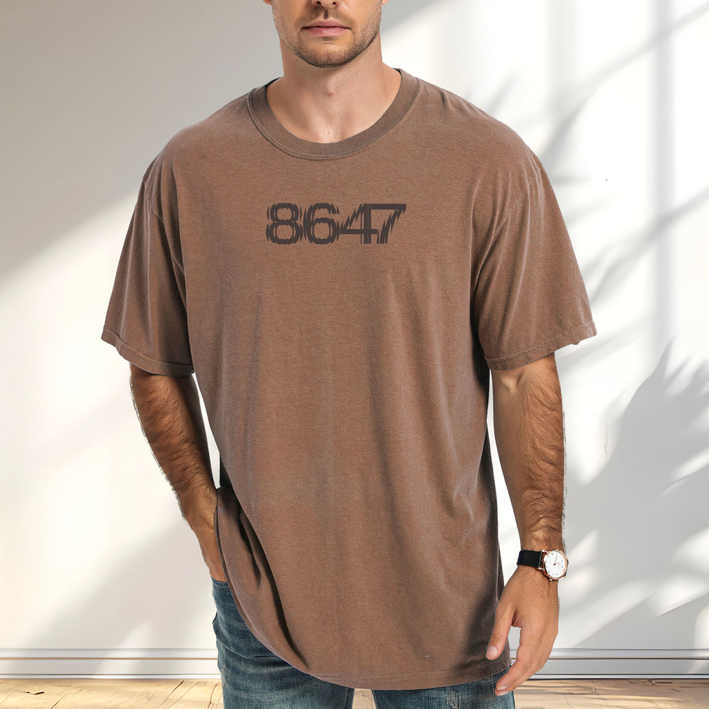 Male model wearing espresso unisex t-shirt featuring the number “8647” in bold, clean typography with glass block effect, referencing Trump impeachment.
