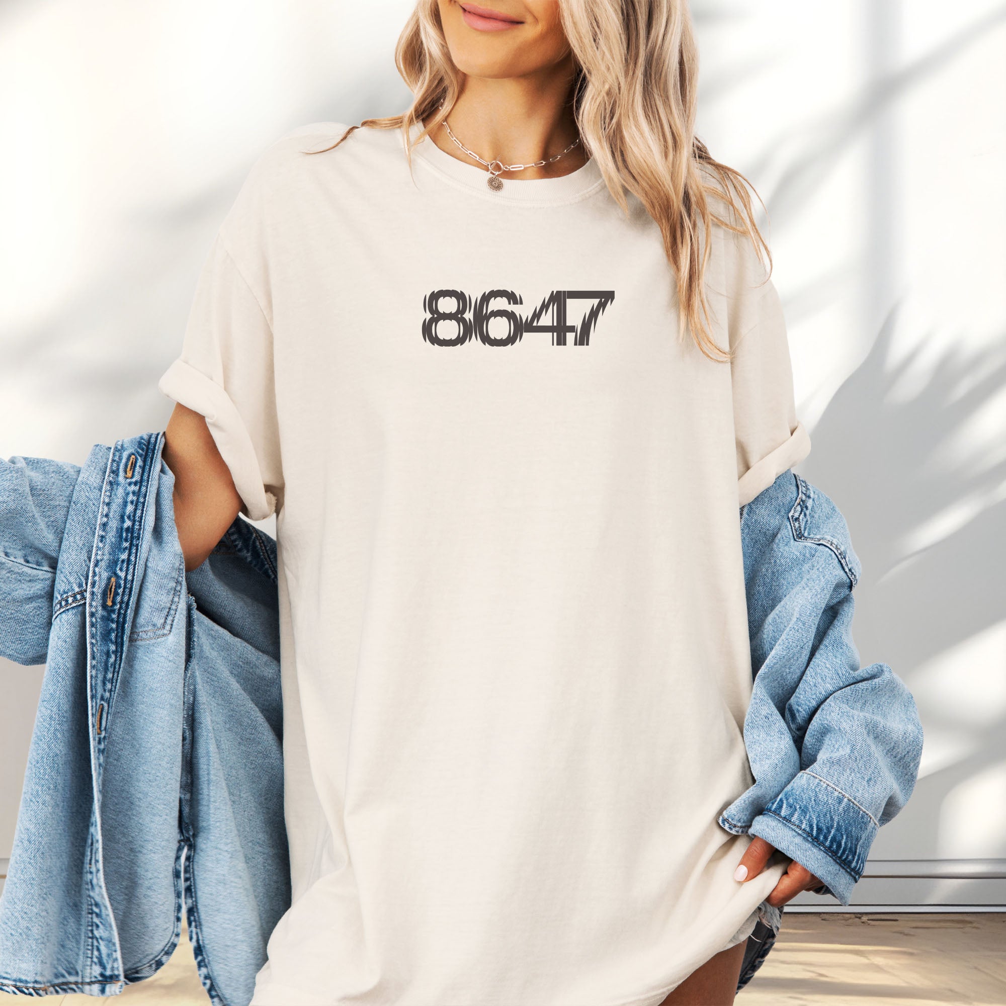Female model wearing oversized ivory unisex t-shirt featuring the number “8647” in bold, clean typography with glass block effect, referencing Trump impeachment.
