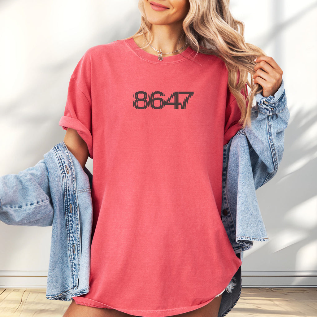 Female model wearing oversized watermelon red unisex t-shirt featuring the number “8647” in bold, clean typography with glass block effect, referencing Trump impeachment.
