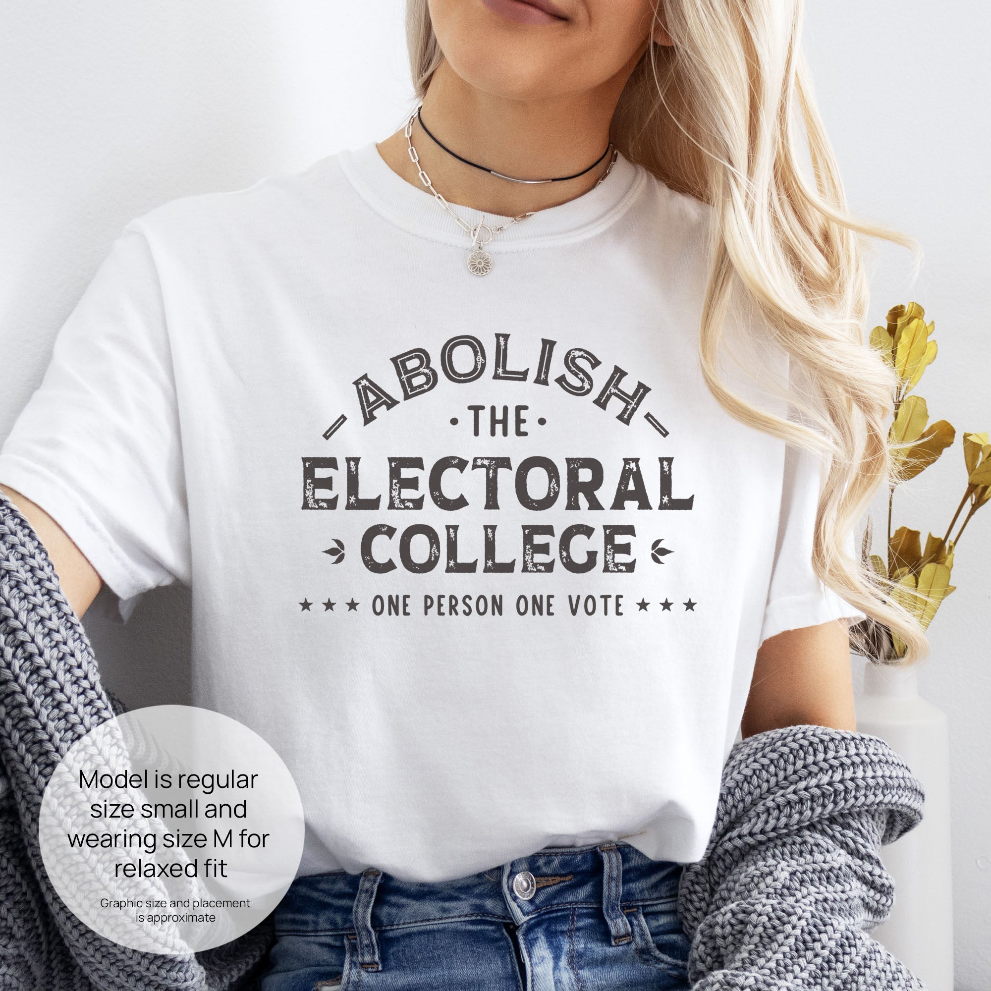Female model wearing white t-shirt with distressed text reading “Abolish the Electoral College One Person One Vote” in vintage style design. Includes size and fit information for the model who is regular size small, and wearing a size M shirt for a relaxed fit.

