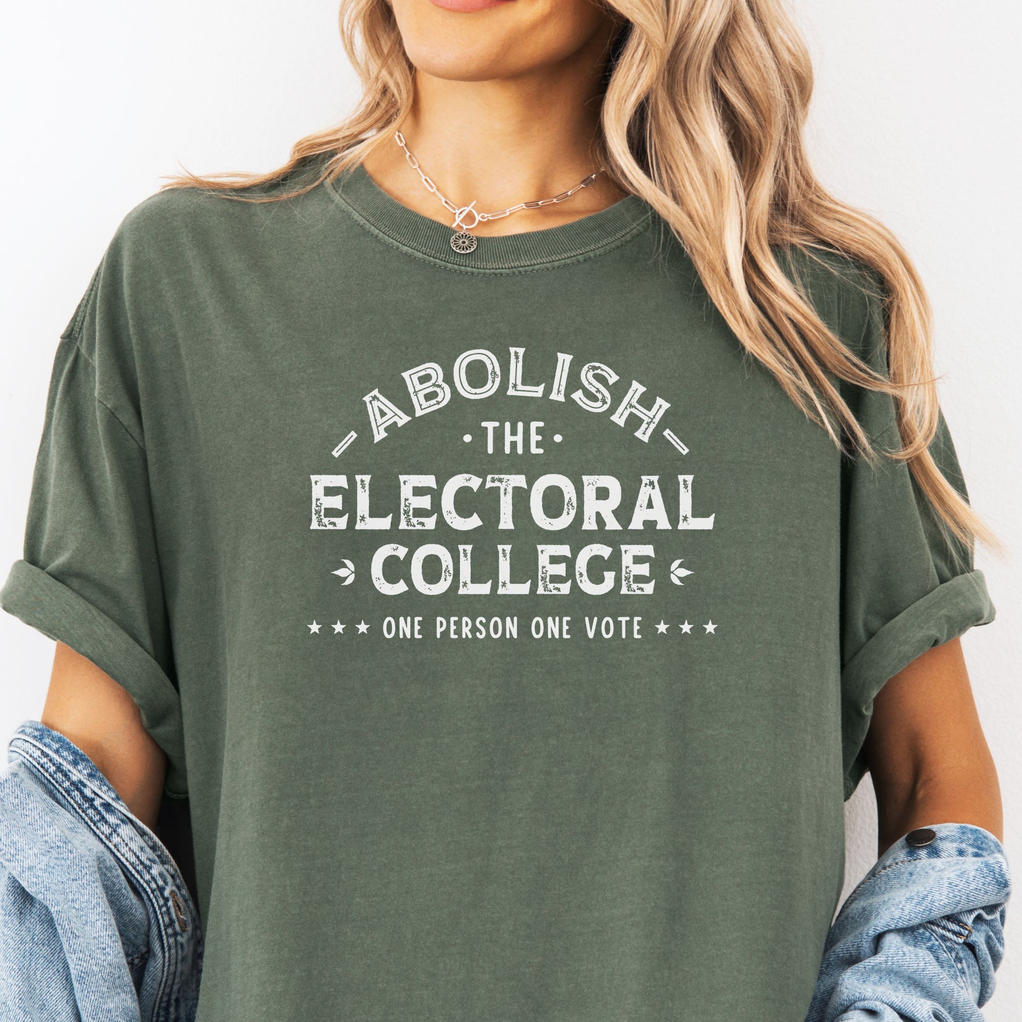 Female model wearing moss Comfort Colors t-shirt with distressed text reading “Abolish the Electoral College One Person One Vote” in vintage style design