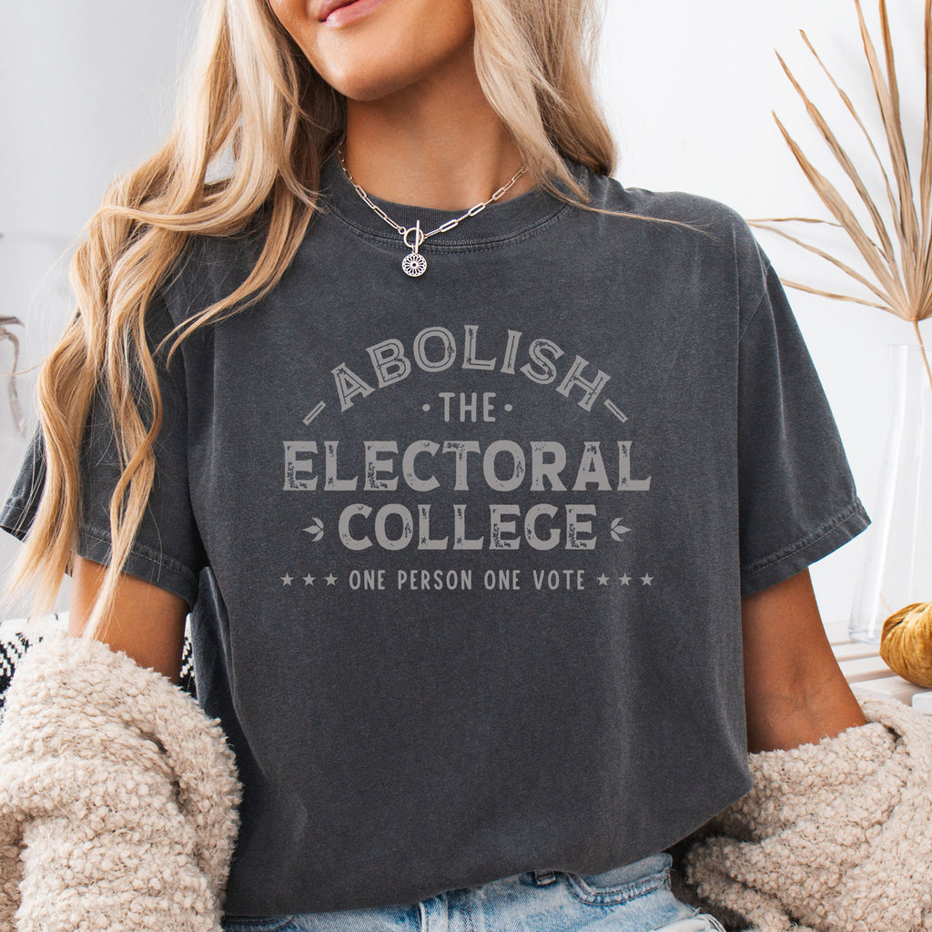 Female model wearing pepper Comfort Colors t-shirt with distressed text reading “Abolish the Electoral College One Person One Vote” in vintage style design