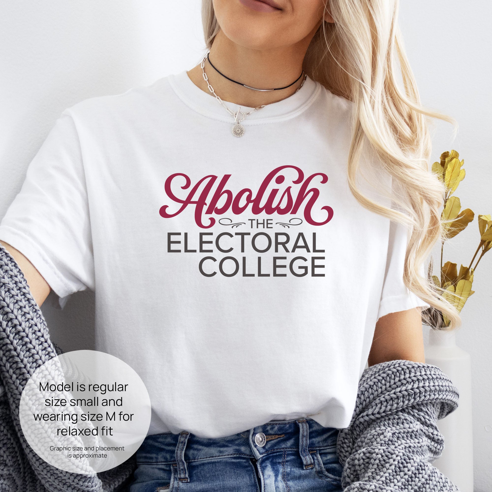 Female model wearing white t-shirt with stylized text reading “Abolish the Electoral College” in muted red and gray. Includes size and fit information for the model who is regular size small, and wearing a size M shirt for a relaxed fit.
