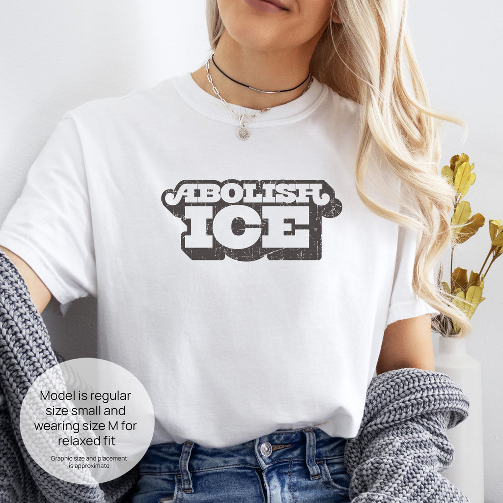 Female model wearing white t-shirt with retro distressed text reading “Abolish ICE” for immigration justice and anti-ICE activism. Includes size and fit information for the model who is regular size small, and wearing a size M shirt for a relaxed fit.
