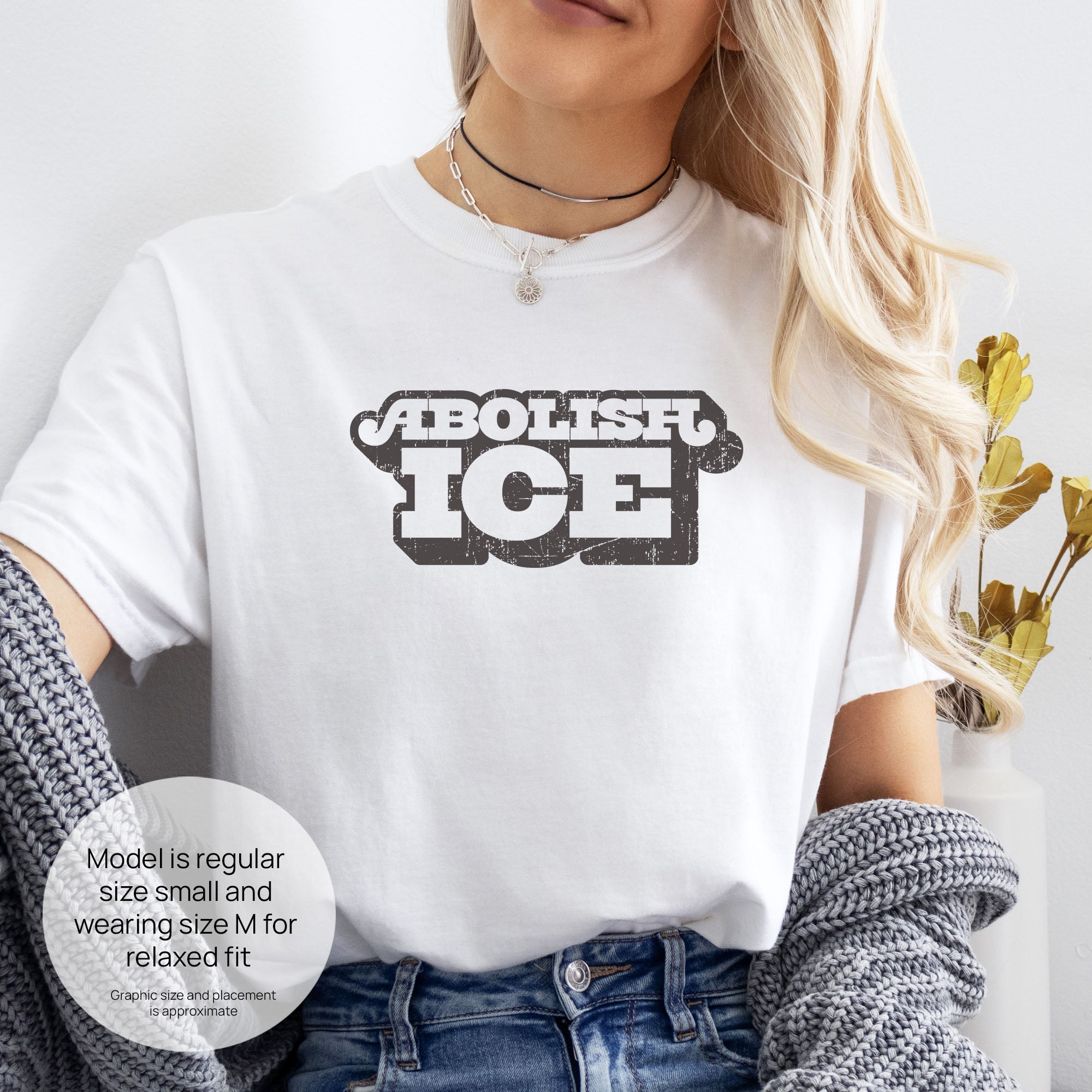 Female model wearing white t-shirt with retro distressed text reading “Abolish ICE” for immigration justice and anti-ICE activism. Includes size and fit information for the model who is regular size small, and wearing a size M shirt for a relaxed fit.
