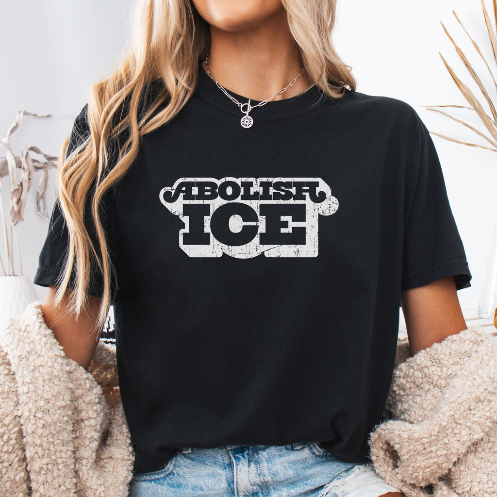 Female model wearing black t-shirt with retro distressed text reading “Abolish ICE” for immigration justice and anti-ICE activism.