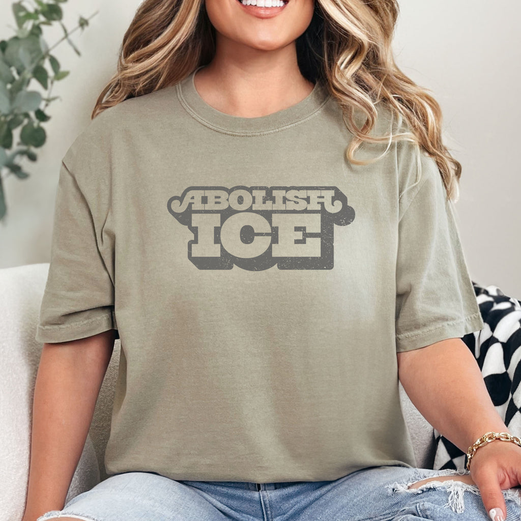 Female model wearing khaki t-shirt with retro text reading “Abolish ICE” for immigration justice and anti-ICE activism.