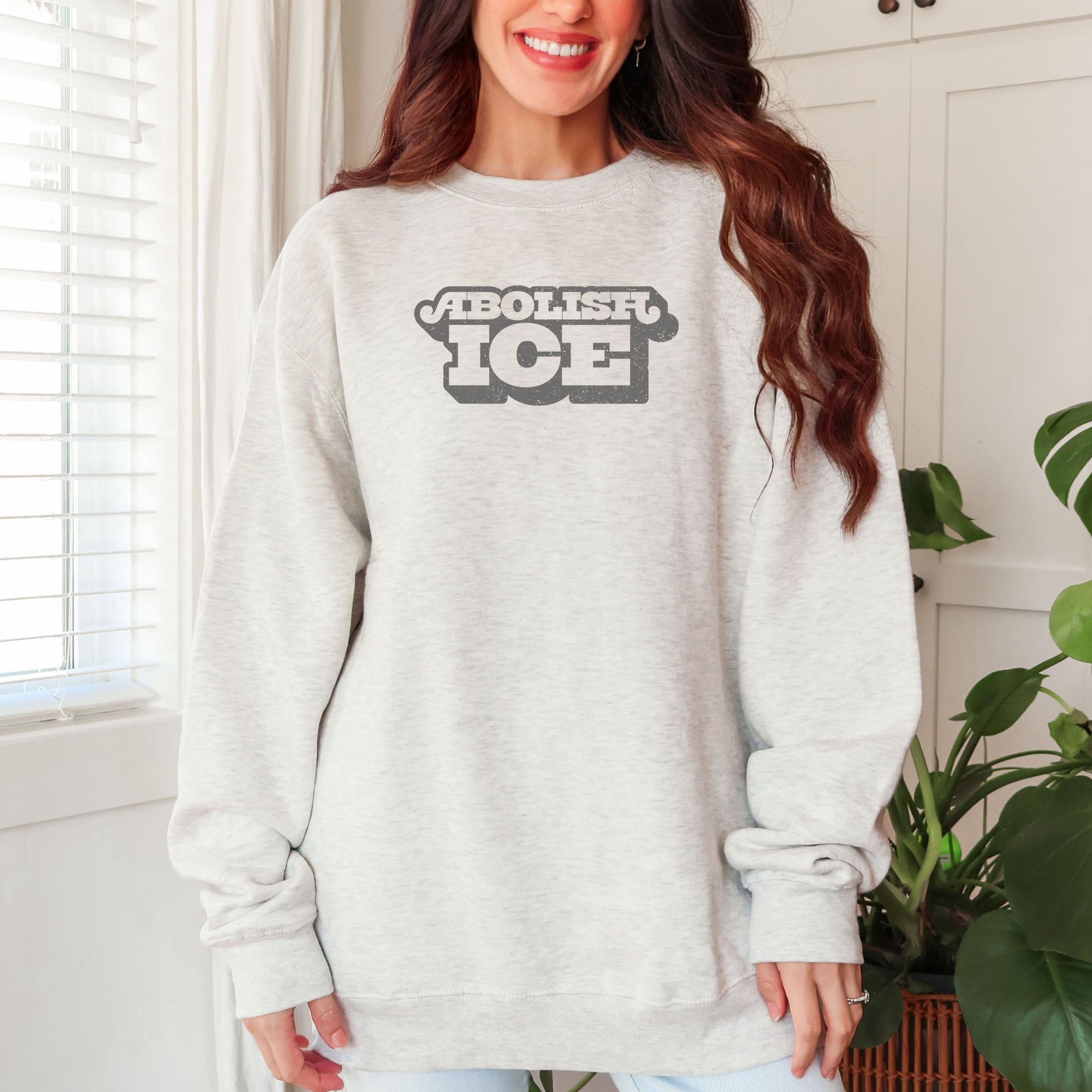 Female model wearing oatmeal sweatshirt with retro text reading “Abolish ICE” for immigration justice and anti-ICE activism.