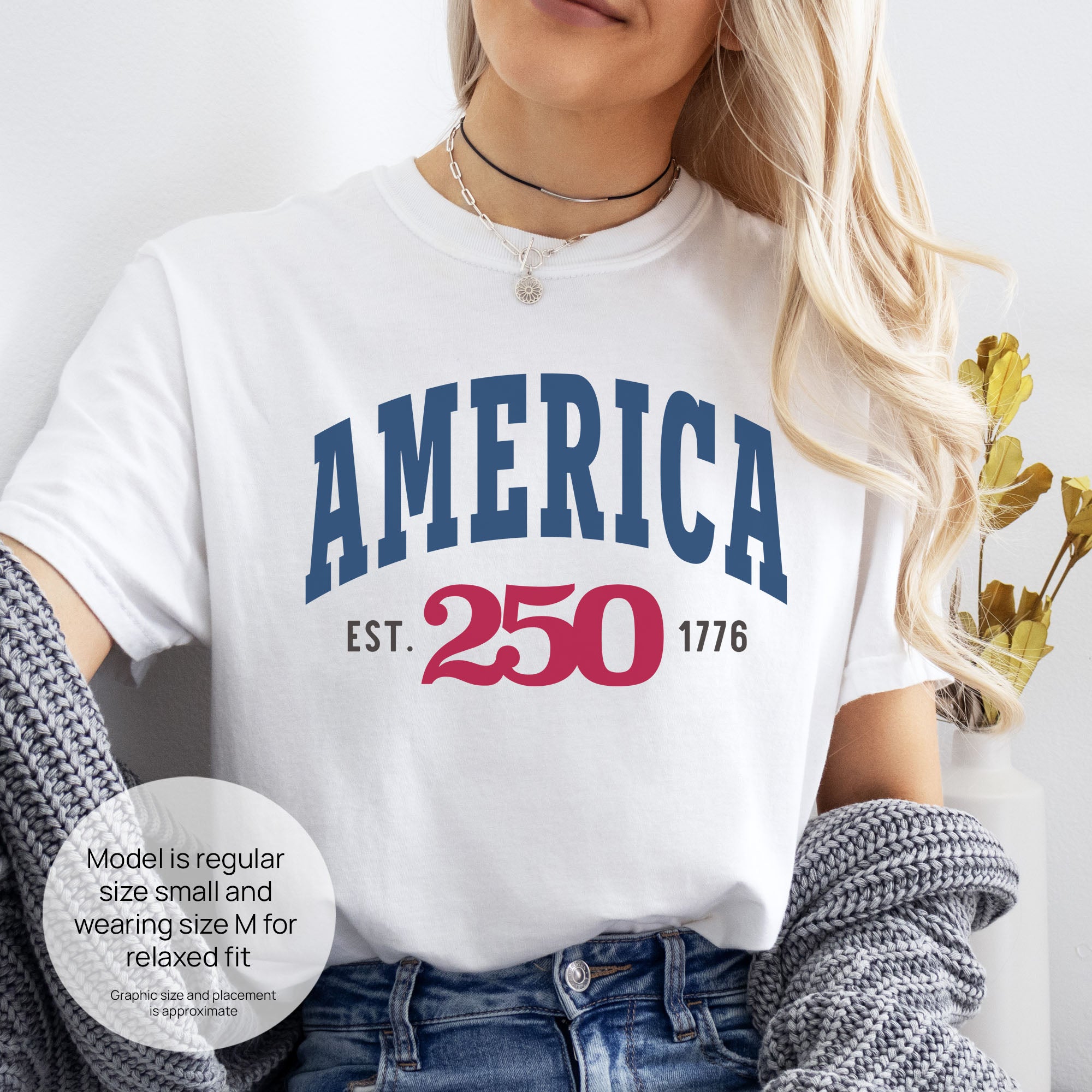 Female model wearing white t-shirt with bold varsity text reading “America 250 — Est. 1776” in varsity style design. Includes size and fit information for the model who is regular size small, and wearing a size M shirt for a relaxed fit.
