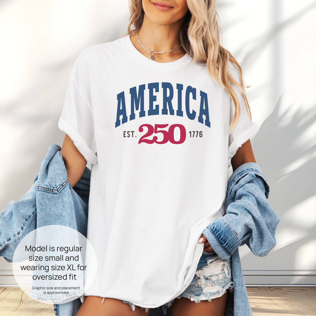 Female model wearing oversized white t-shirt with bold varsity text reading “America 250 — Est. 1776” in varsity style design. Includes size and fit information for the model who is regular size small, and wearing a size XL shirt for an oversized fit.
