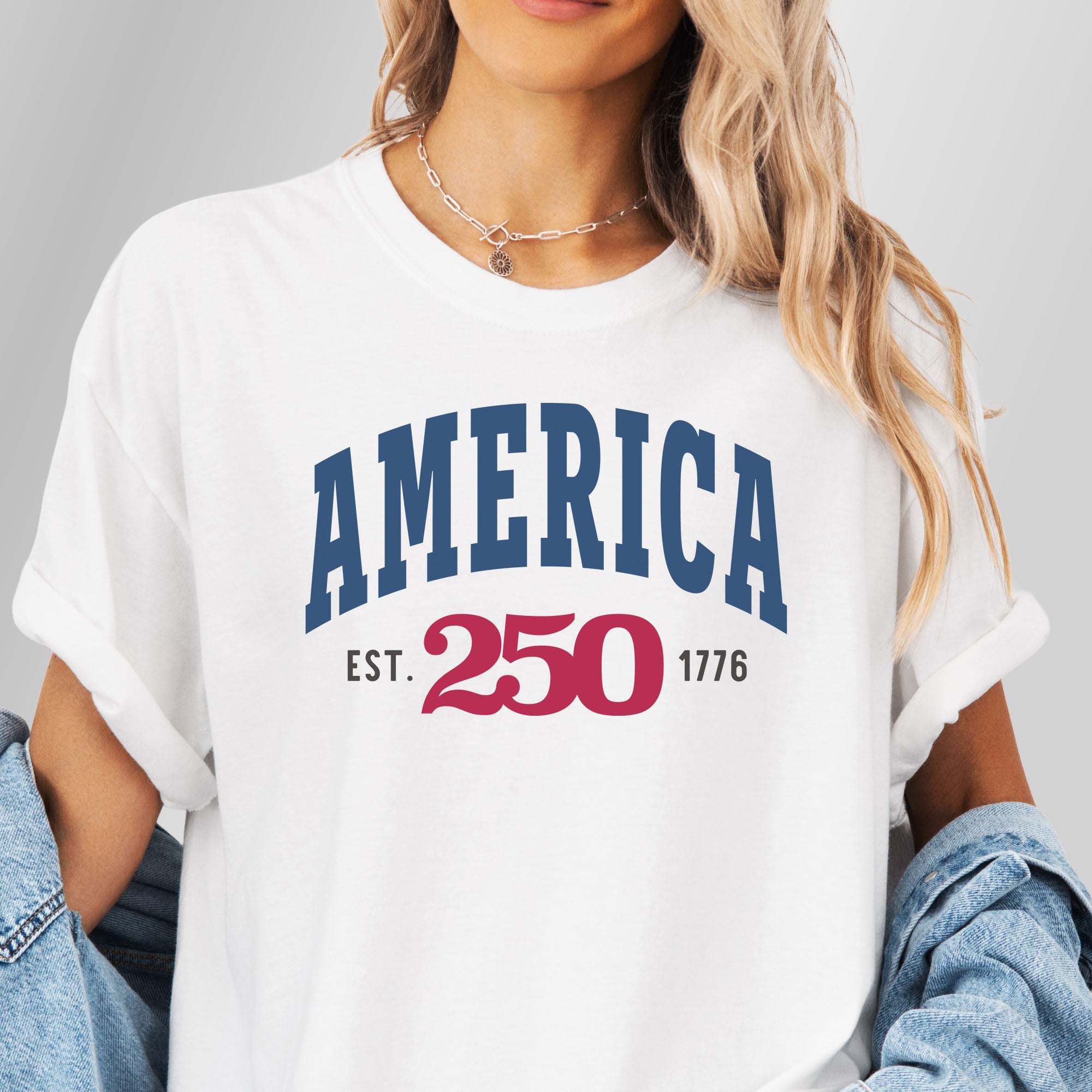Female model wearing white t-shirt with bold varsity text reading “America 250 — Est. 1776” in varsity style design.