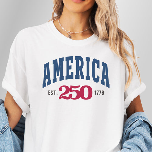 Female model wearing white t-shirt with bold varsity text reading “America 250 — Est. 1776” in varsity style design.