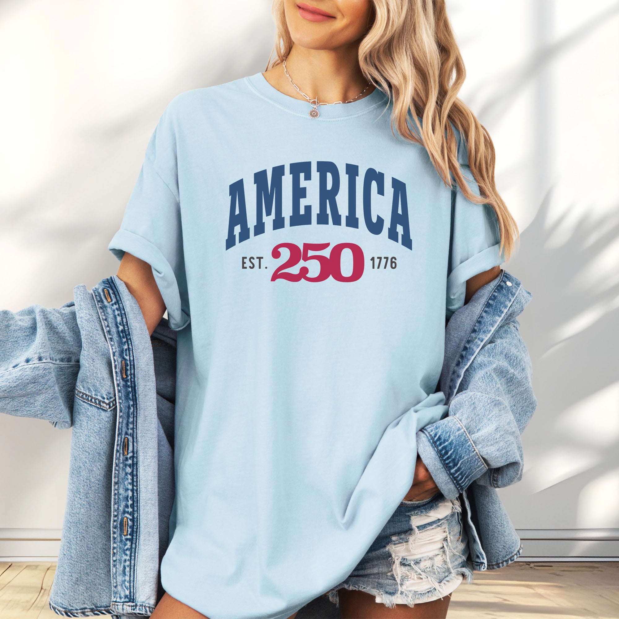 Female model wearing oversized chambray t-shirt with bold varsity text reading “America 250 — Est. 1776” in varsity style design.