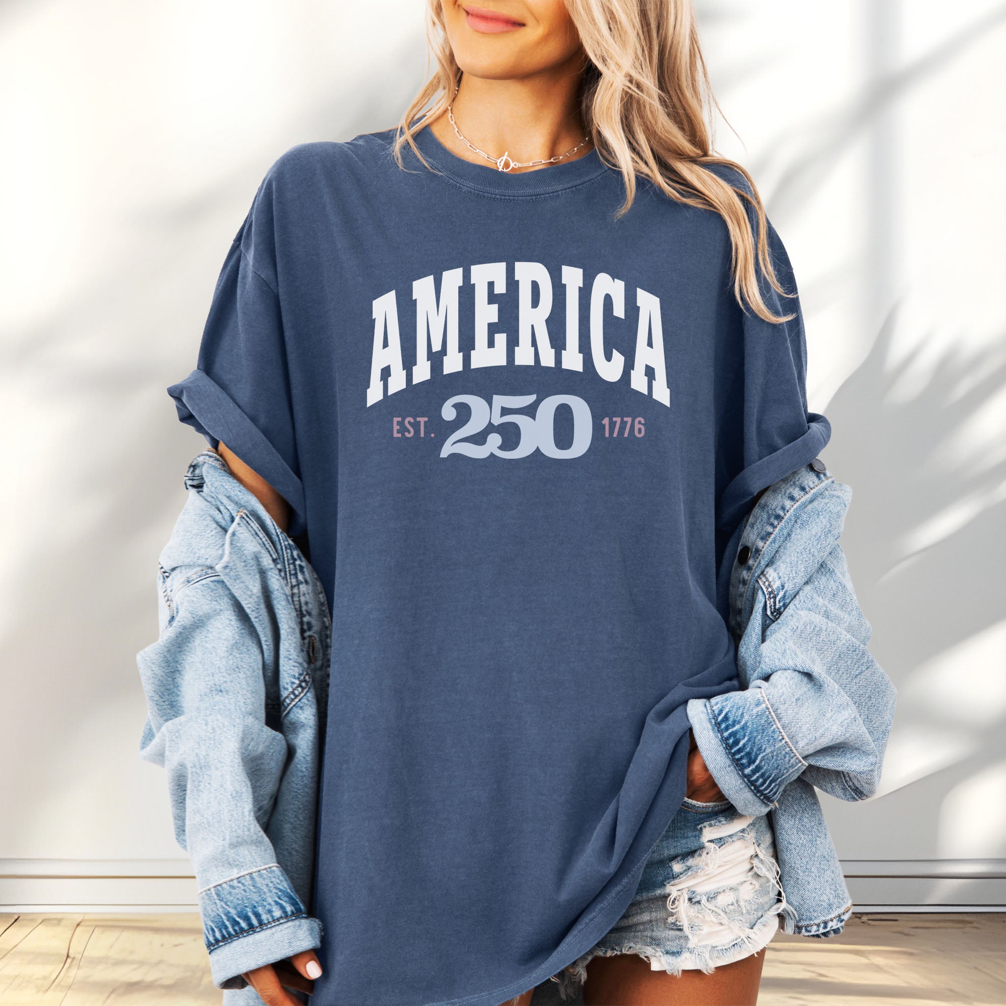 Female model wearing oversized denim t-shirt with bold varsity text reading “America 250 — Est. 1776” in varsity style design.