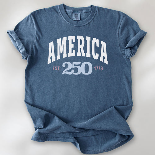 Denim t-shirt with bold varsity text reading “America 250 — Est. 1776” in varsity style design. 