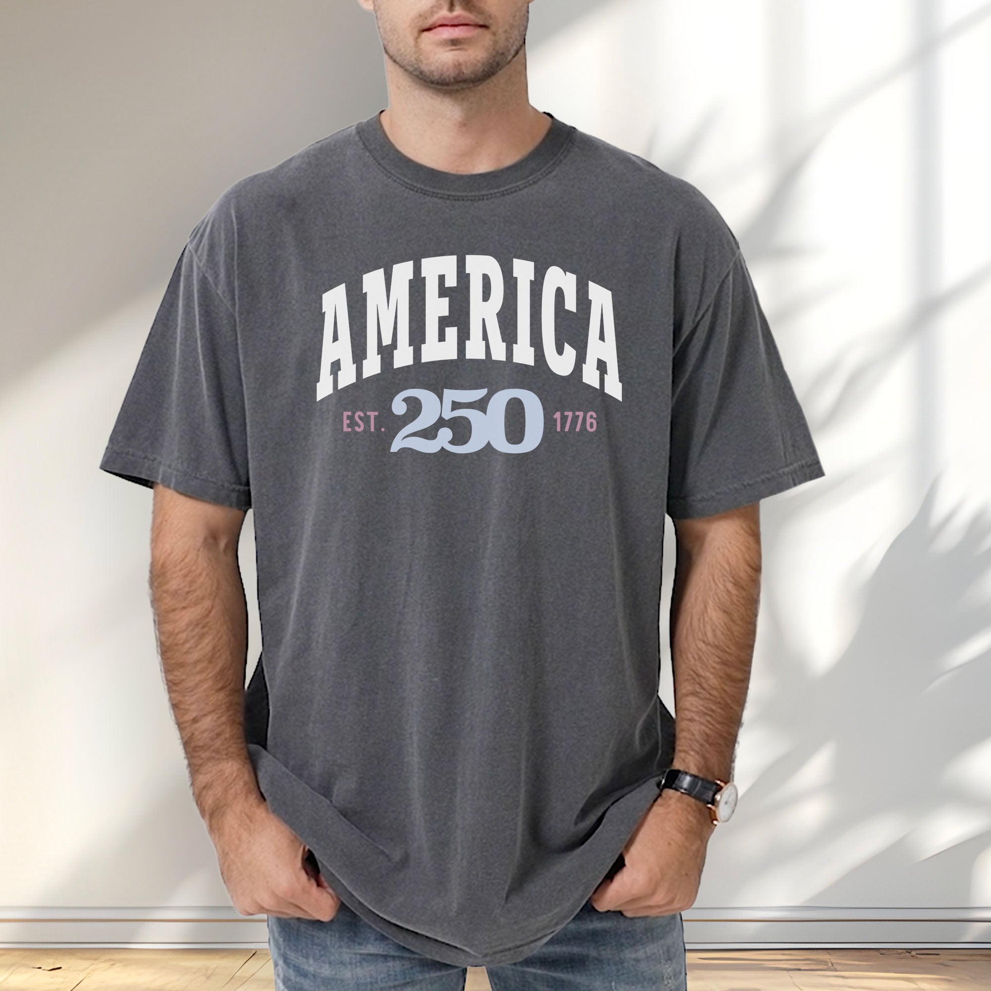 Male model wearing pepper t-shirt with bold varsity text reading “America 250 — Est. 1776” in varsity style design.
