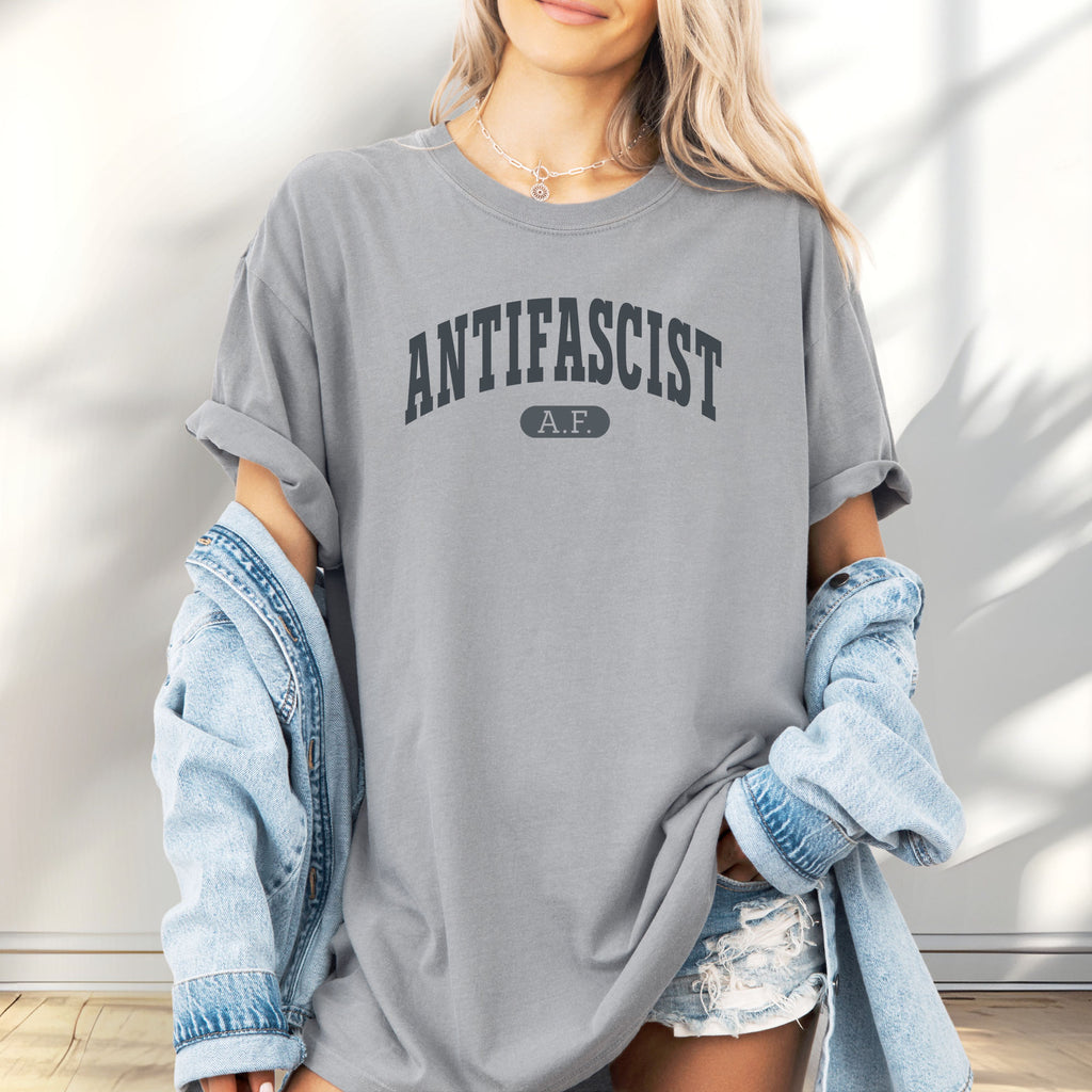 Female model wearing granite Comfort Colors t-shirt with varsity collegiate style text across the front that reads "Antifascist AF."