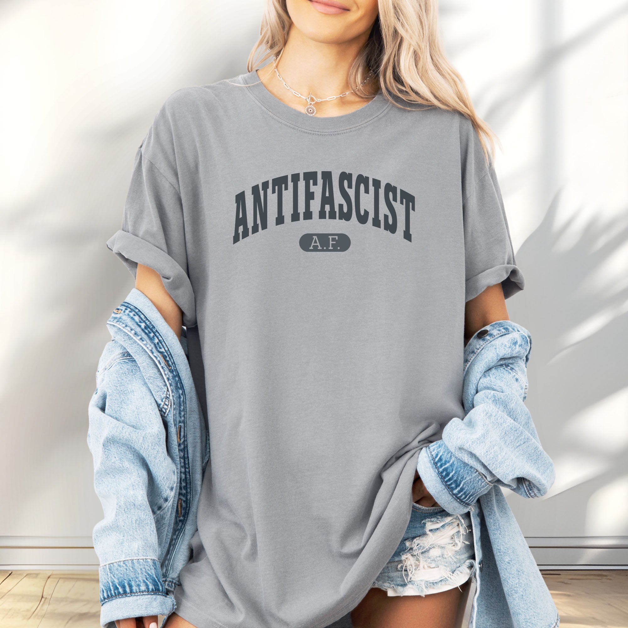 Female model wearing granite Comfort Colors t-shirt with varsity collegiate style text across the front that reads "Antifascist AF."