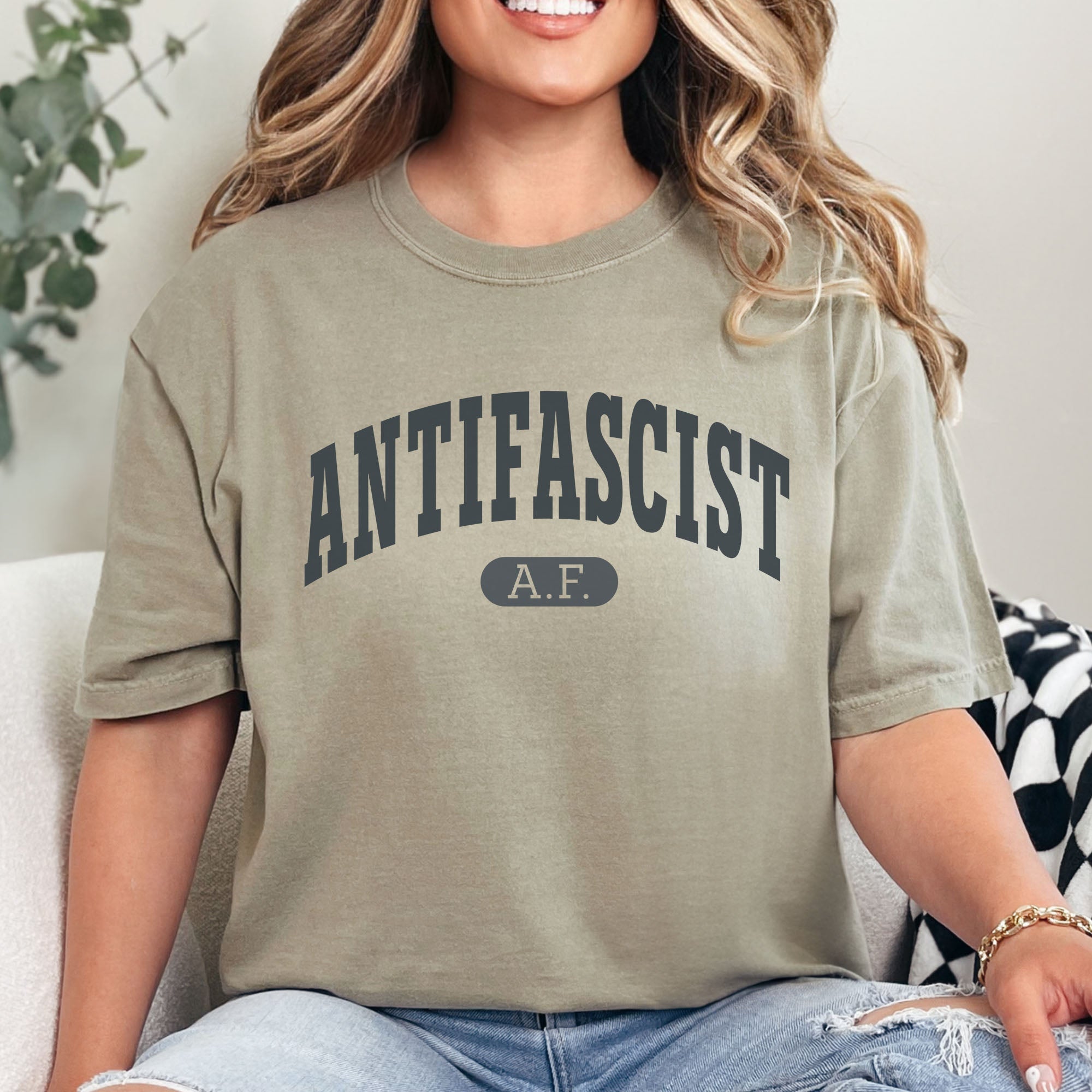 Female model wearing khaki Comfort Colors t-shirt with varsity collegiate style text across the front that reads "Antifascist AF."