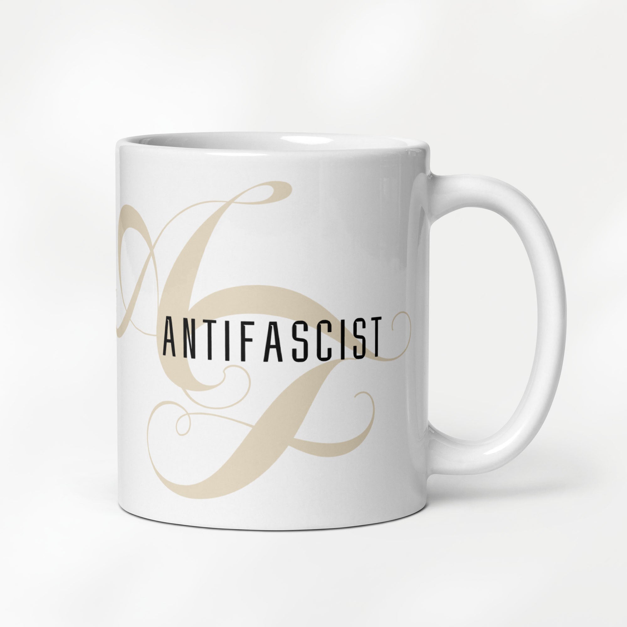 11 ounce white ceramic mug with the word “Antifascist” in black text and a subtle light tan script design of the letters A and F in the background.