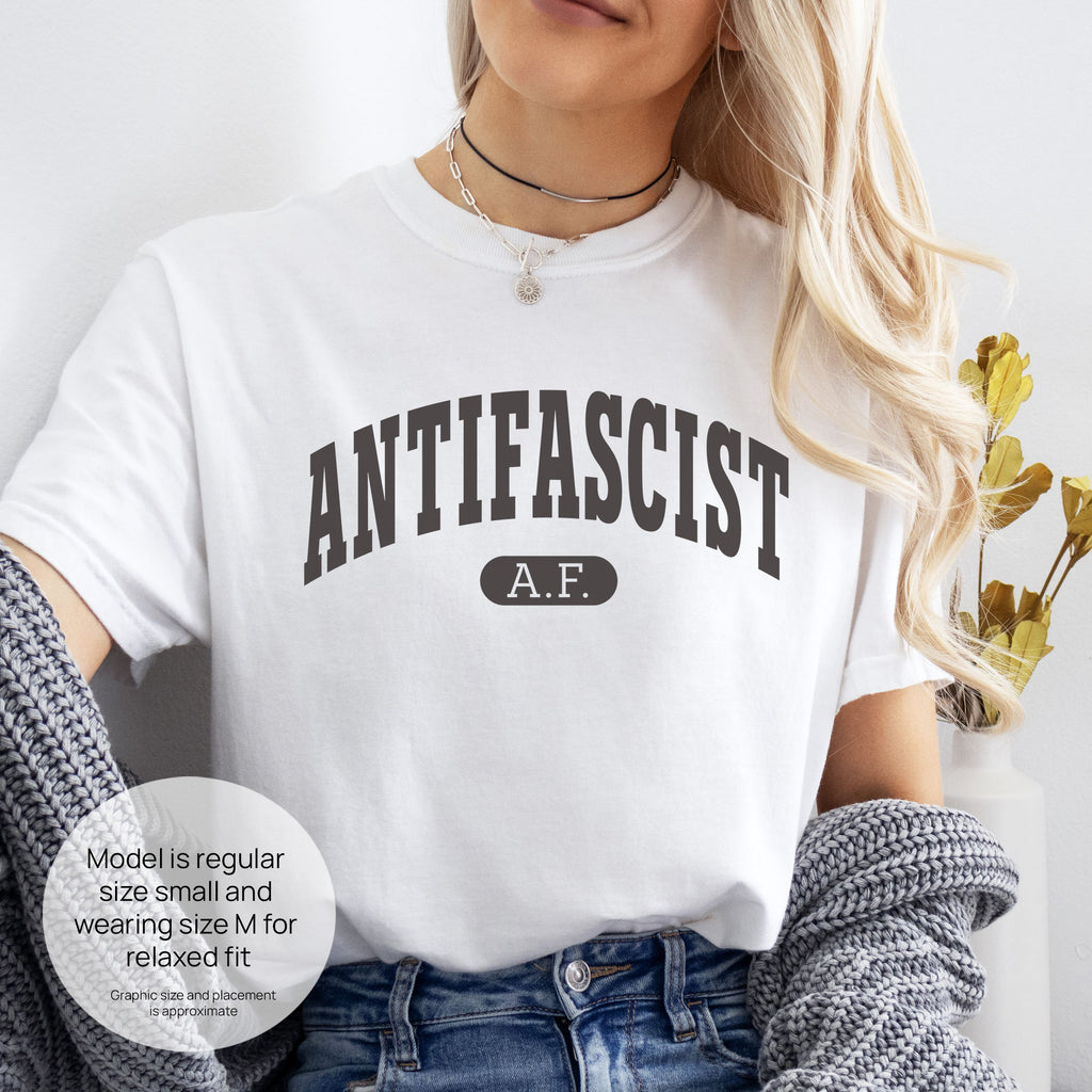 Female model wearing white Comfort Colors t-shirt with varsity collegiate style text across the front that reads "Antifascist AF." Includes size and fit information for the model who is regular size small, and wearing a size M shirt for a relaxed fit.
