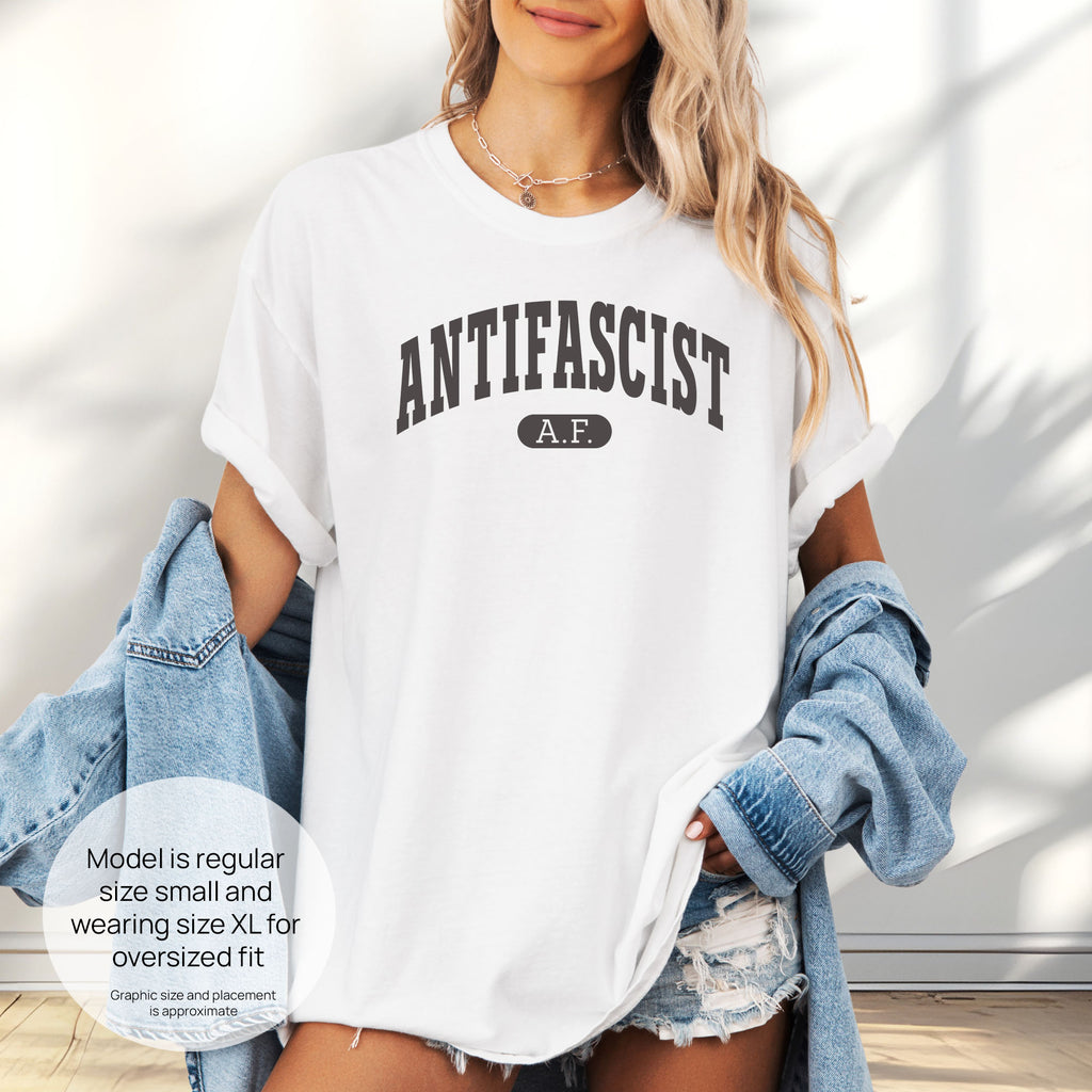 Female model wearing white Comfort Colors t-shirt with varsity collegiate style text across the front that reads "Antifascist AF." Includes size and fit information for the model who is regular size small, and wearing a size XL shirt for an oversized fit.
