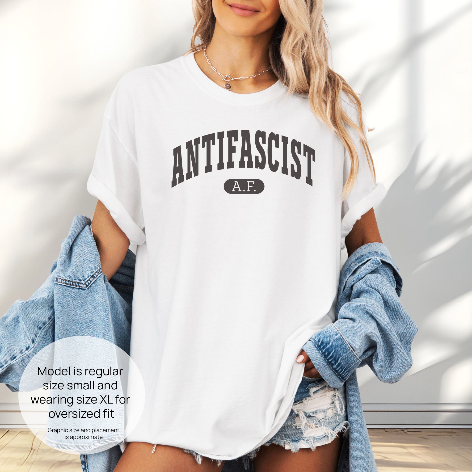 Female model wearing white Comfort Colors t-shirt with varsity collegiate style text across the front that reads "Antifascist AF." Includes size and fit information for the model who is regular size small, and wearing a size XL shirt for an oversized fit.

