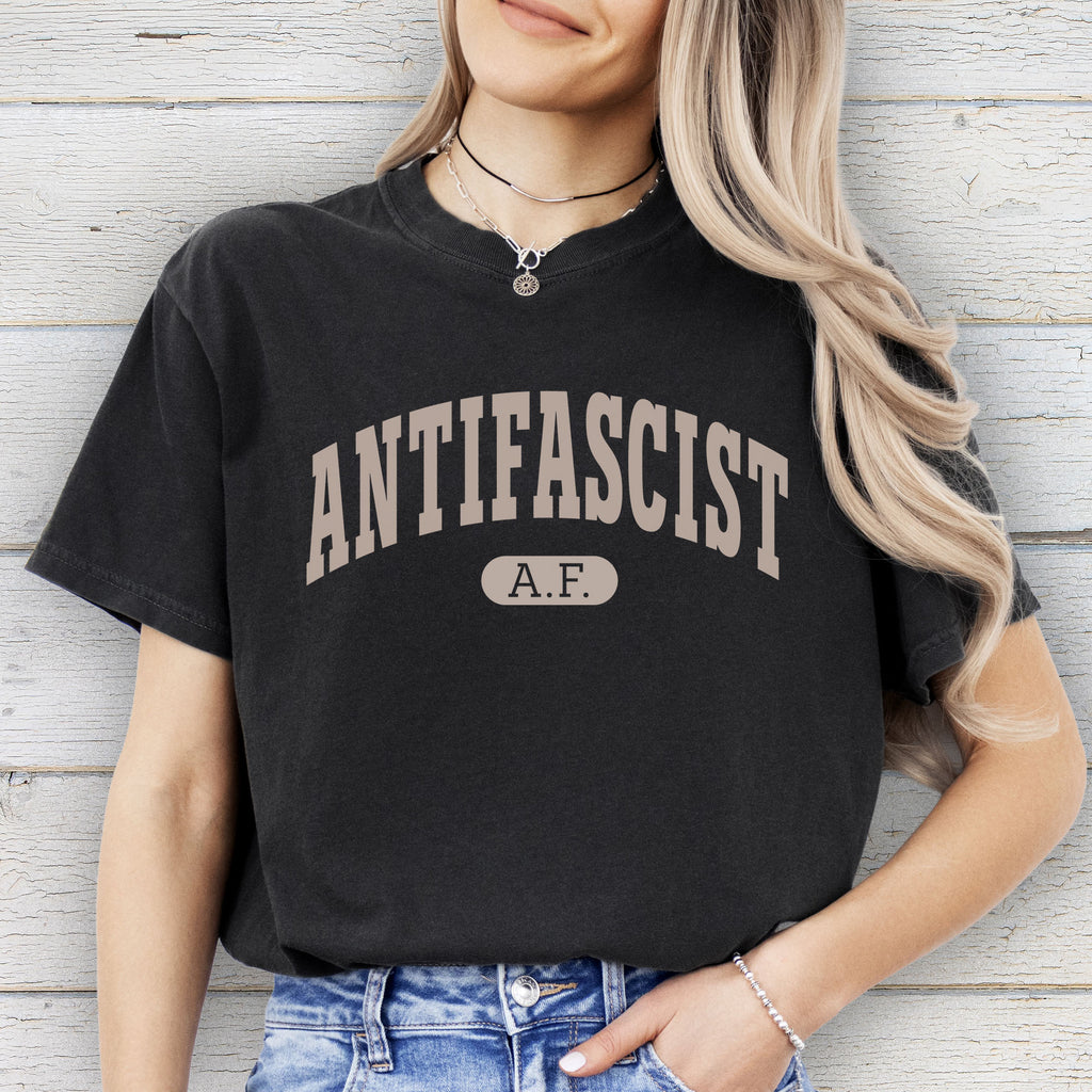 Female model wearing black Comfort Colors t-shirt with varsity collegiate style text across the front that reads "Antifascist AF."