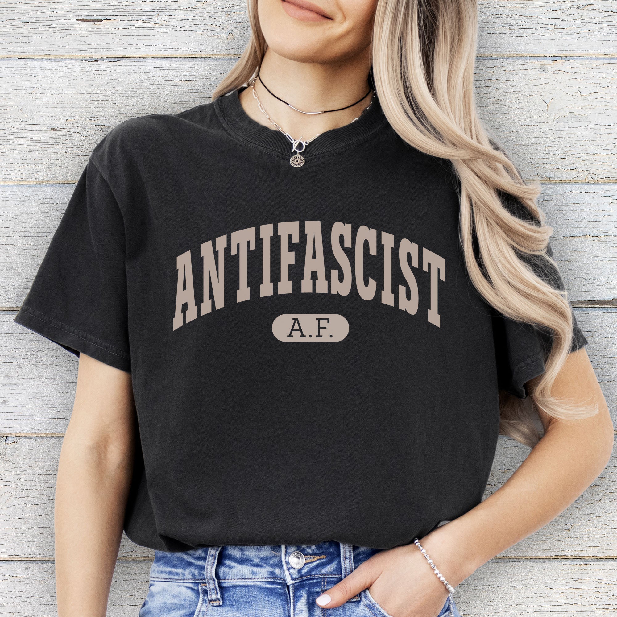 Female model wearing black Comfort Colors t-shirt with varsity collegiate style text across the front that reads "Antifascist AF."