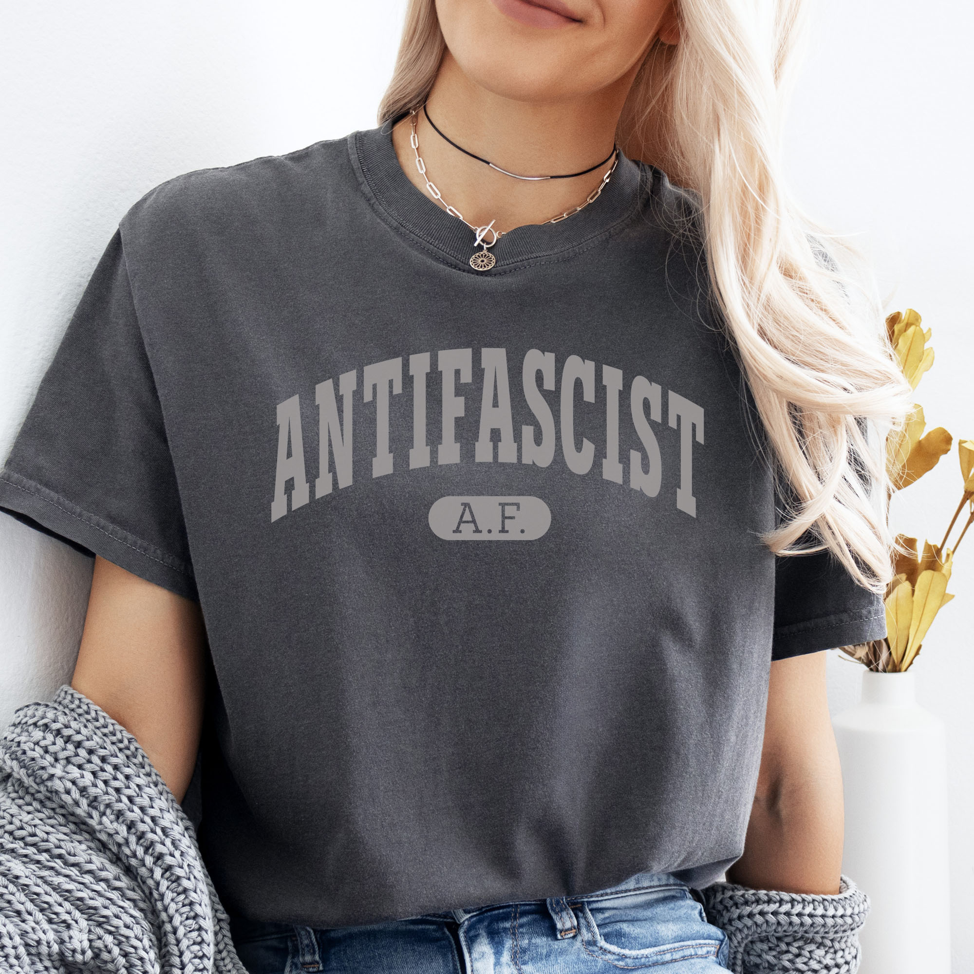 Female model wearing pepper Comfort Colors t-shirt with varsity collegiate style text across the front that reads "Antifascist AF."