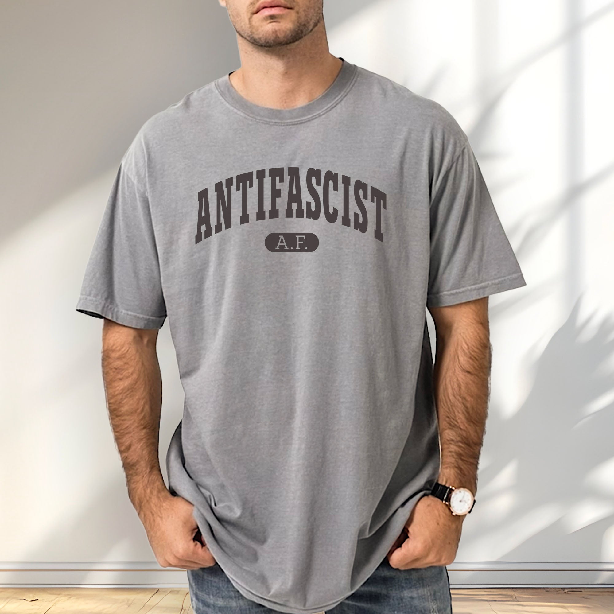 Male model wearing grey Comfort Colors t-shirt with varsity collegiate style text across the front that reads "Antifascist AF."