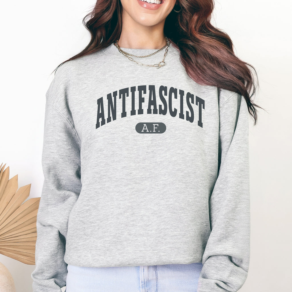 Female model wearing grey crewneck sweatshirt with varsity collegiate style text across the front that reads "Antifascist AF."