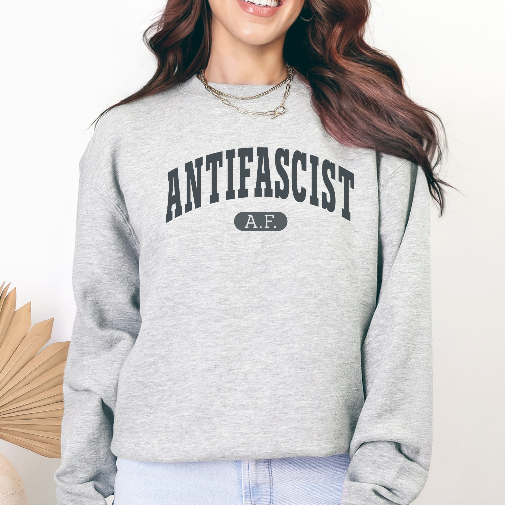 Female model wearing grey crewneck sweatshirt with varsity collegiate style text across the front that reads "Antifascist AF."
