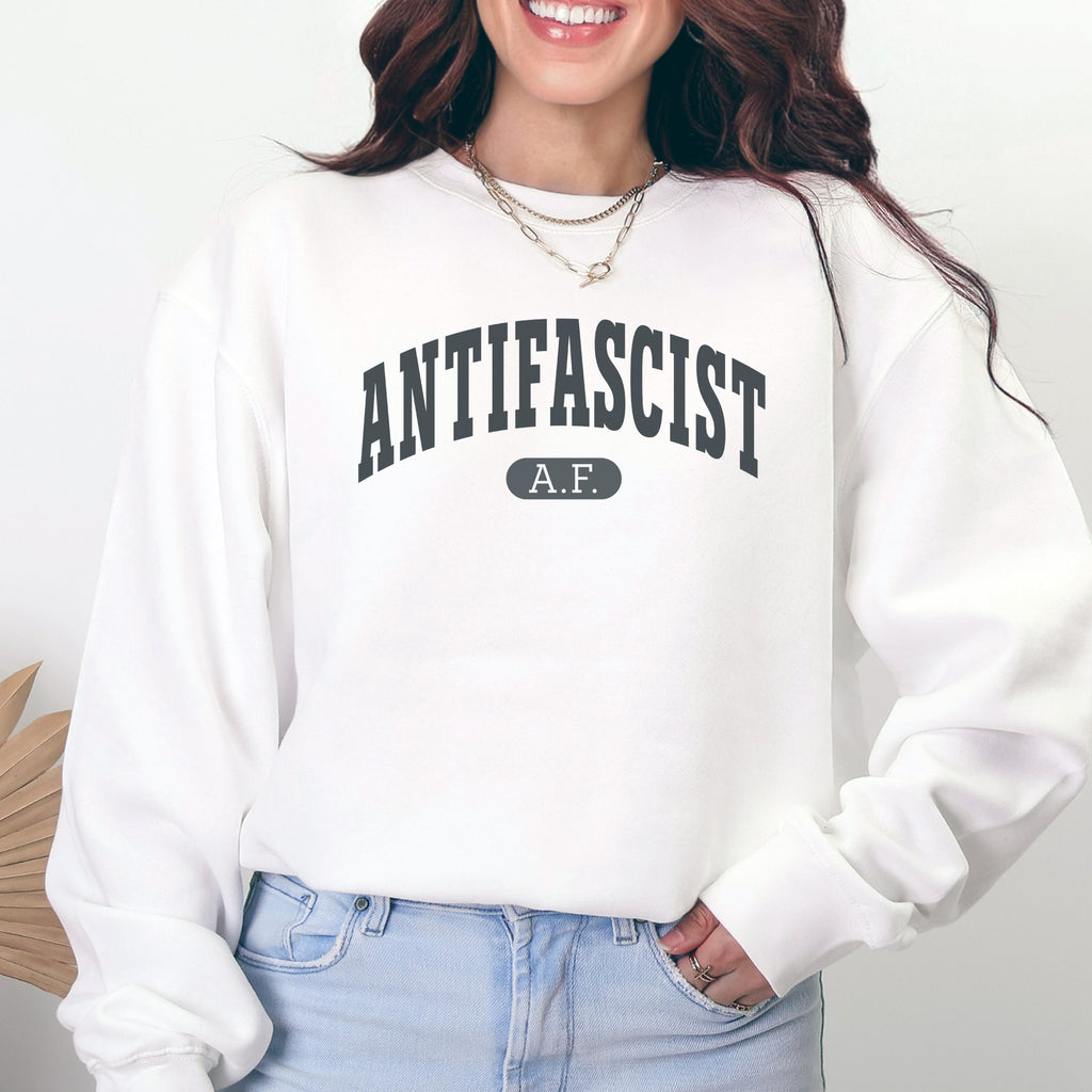 Female model wearing white crewneck sweatshirt with varsity collegiate style text across the front that reads "Antifascist AF."