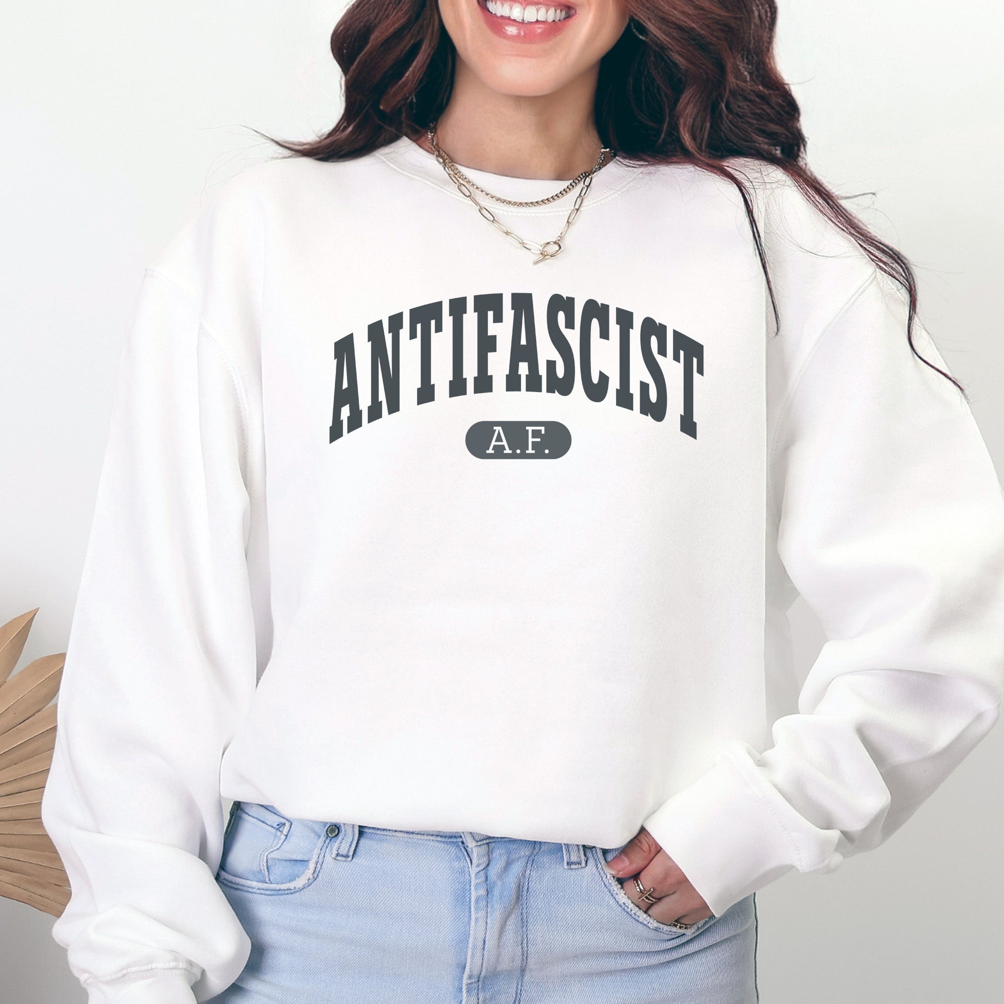Female model wearing white crewneck sweatshirt with varsity collegiate style text across the front that reads "Antifascist AF."