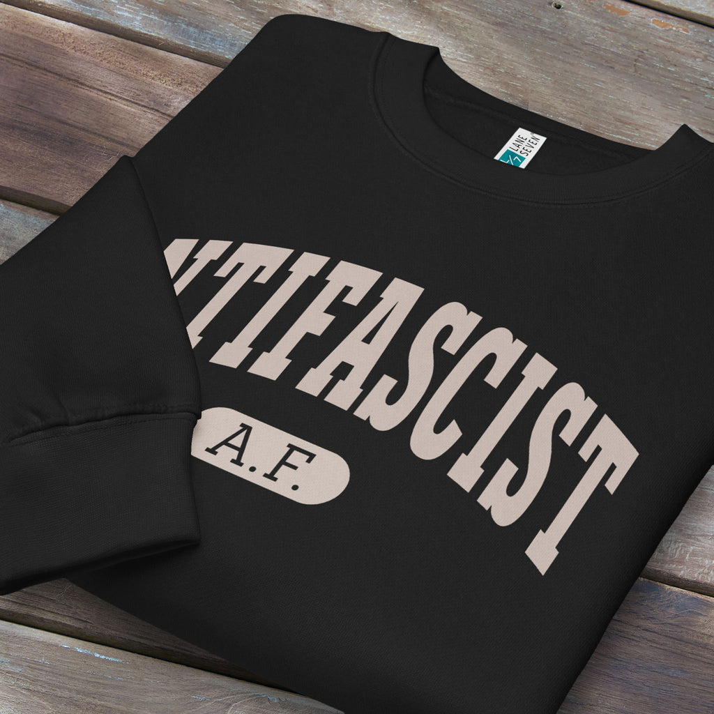 Folded black crewneck sweatshirt with varsity collegiate style text across the front that reads "Antifascist AF."