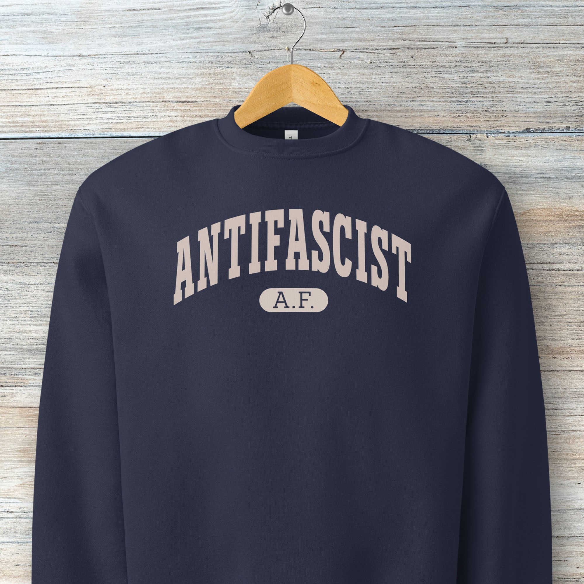 Hanging navy Bella + Canvas heavyweight crewneck sweatshirt with varsity collegiate style text across the front that reads "Antifascist AF."