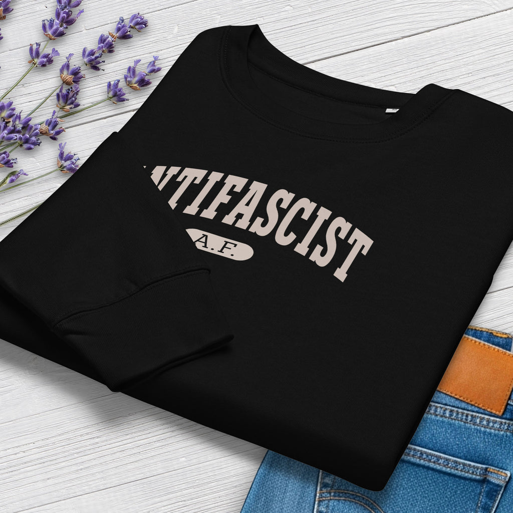 Folded black sweatshirt with greyish tan text that reads “Antifascist A.F.” in a collegiate style. Shows closeup cuff detail, styled with greenery and jeans.