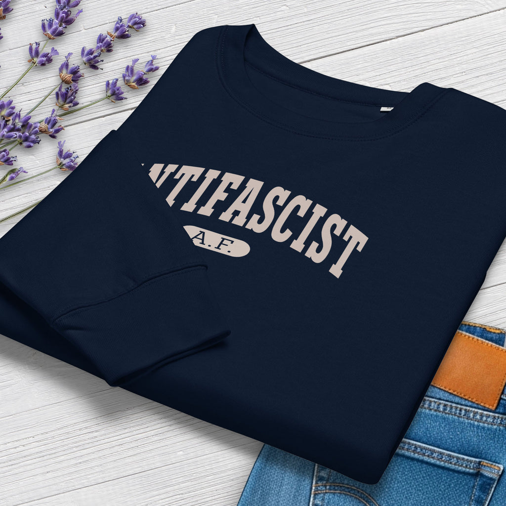 Folded navy sweatshirt with greyish tan text that reads “Antifascist A.F.” in a collegiate style. Shows closeup cuff detail, styled with greenery and jeans.